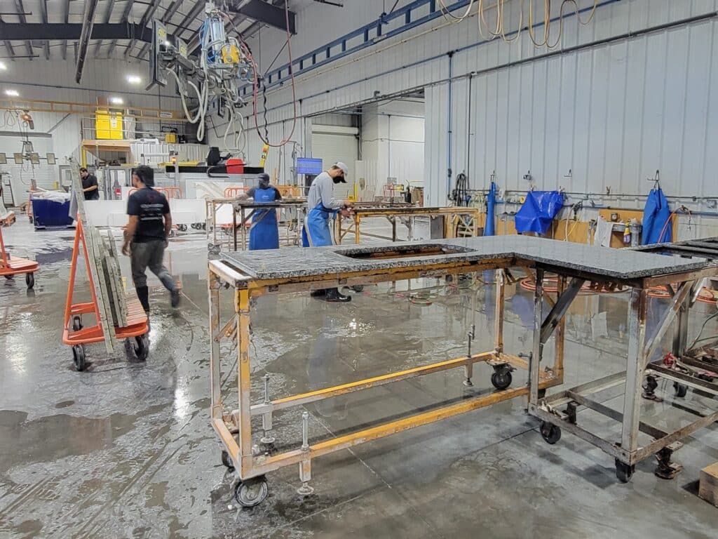 Shop - Stone Fabricator Solutions - Stone Fabricator Solutions Industrial workshop with workers in blue aprons and hard hats handling granite slabs on wheeled carts; wet floor and overhead machinery visible in the background.