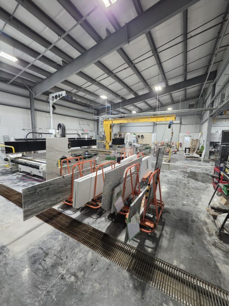 20260501_075617 - Stone Fabricator Solutions - Stone Fabricator Solutions Industrial stone fabrication shop with large granite slabs resting on orange stands beside a metal drainage trench and a yellow overhead crane in the background