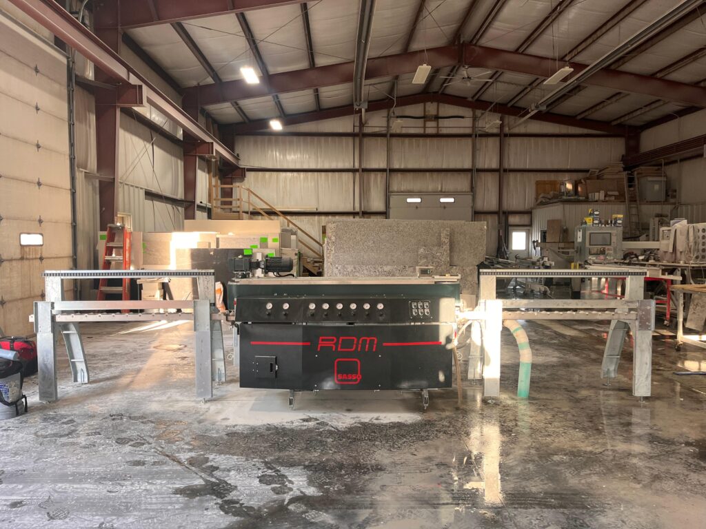 IMG-20260506-WA0018 - Stone Fabricator Solutions - Stone Fabricator Solutions Industrial workshop with a large stone-cutting machine in the foreground and granite slabs stacked along walls.
