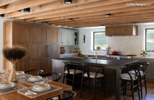 Kitchen - Stone Fabricator Solutions Modern rustic kitchen with a large island, exposed wooden ceiling beams, and a dining table set for six in the foreground.