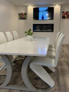Litten 1 - Stone Fabricator Solutions Modern conference room with a long white marble table, white chairs, a potted plant centerpiece, wall-mounted TV, and a fireplace above marble wall panels.