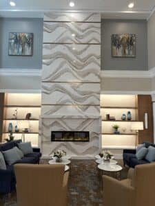 image0 - Stone Fabricator Solutions Tall marble-column fireplace centerpiece with gray veining in a modern living room, flanked by wall art and built-in shelves.