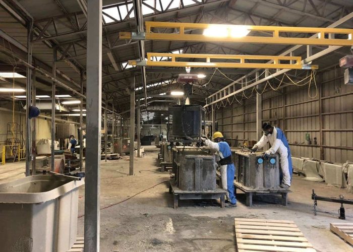 Industrial workshop with two workers in blue overalls and yellow hard hats operating large machinery on concrete blocks under overhead cranes in a spacious warehouse, creating concrete molds.