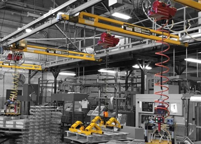 Industrial manufacturing floor with overhead yellow crane rails, red hoists, spiral hoses, and various machines and control panels.