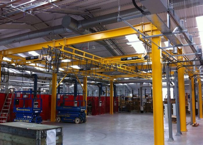 Industrial workshop with yellow overhead crane beams and blue scissor lift platforms inside a factory setting.