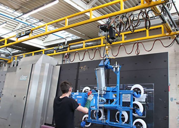 Worker in a black shirt operates a blue, wheeled testing rig inside an industrial facility, with a yellow overhead gantry and large metal cabinets nearby.