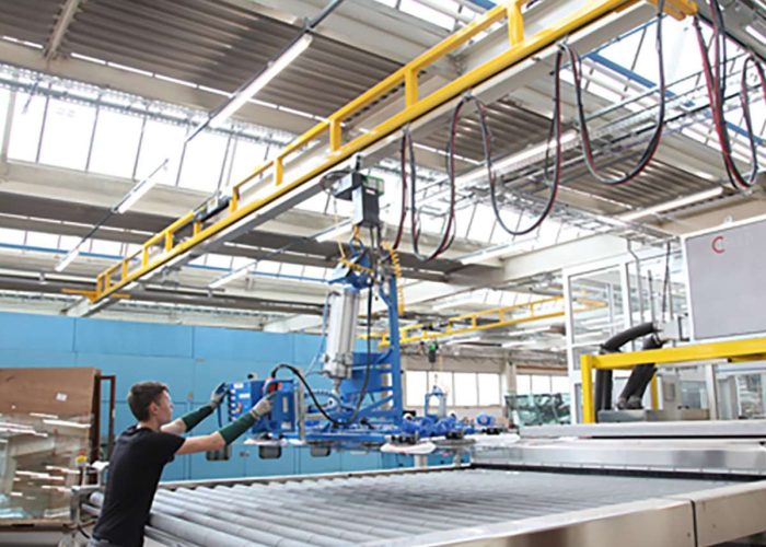 Man in a black shirt operates a blue robotic arm over a metal conveyor in a bright industrial factory setting.