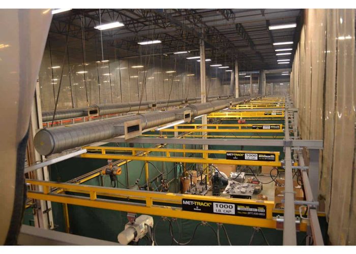 Industrial workshop with yellow overhead crane rails, large ducting along the ceiling and a green floor below.